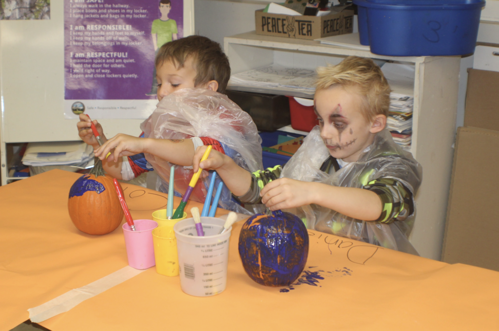 Two children in plastic aprons paint pumpkins bright blue during a classroom art activity.