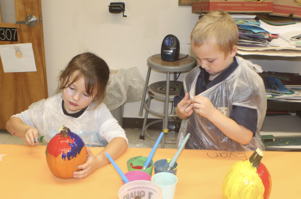 A girl paints a red-and-blue pumpkin while her classmate adjusts his apron beside her.