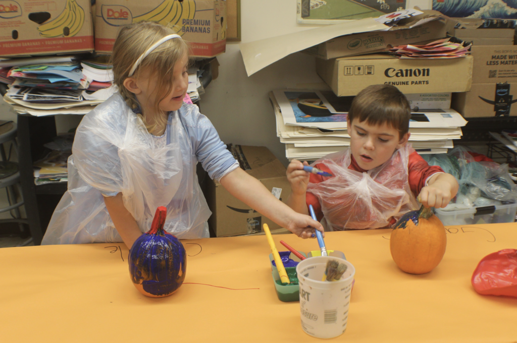 Two students share paintbrushes and laugh while decorating pumpkins with bright colors.