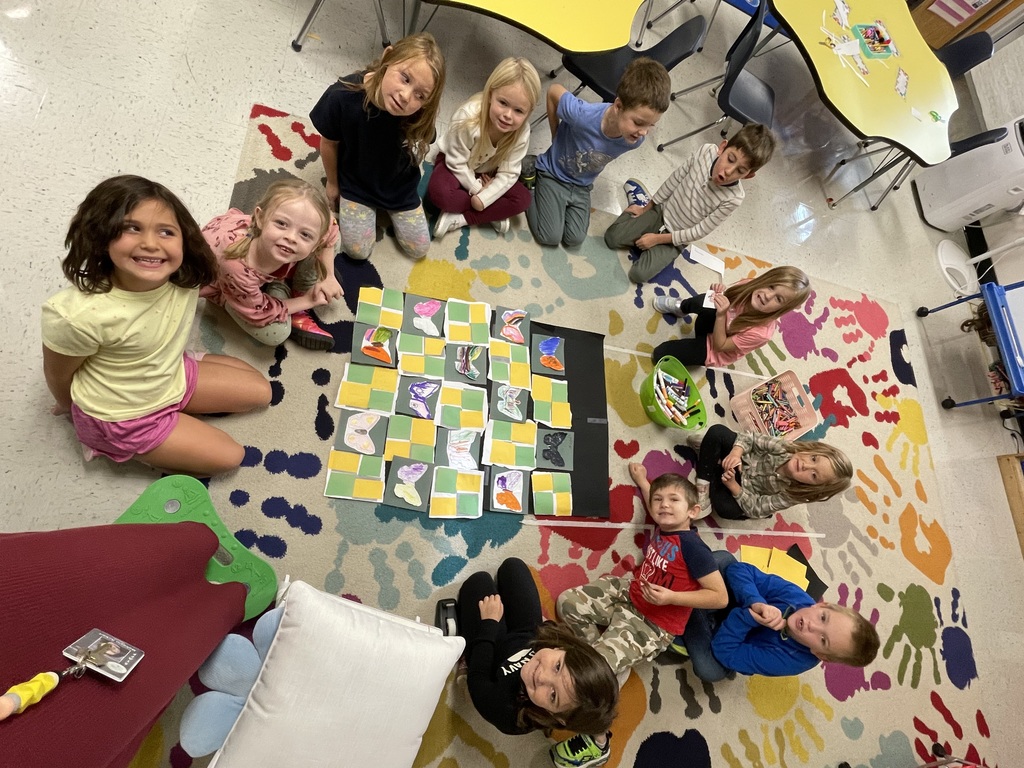 A group of young children sit in a circle on a colorful rug in a classroom, smiling and looking up at the camera. In the center of the group is a project made of yellow and green squares with cut-out, hand-drawn shapes placed on top. Tables with art supplies are visible in the background.