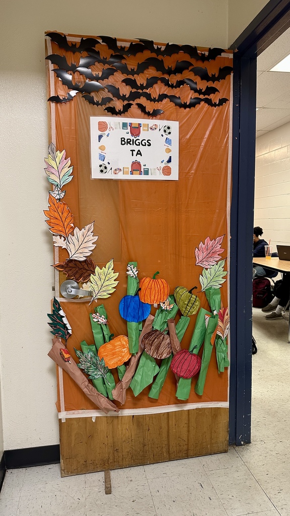 Door decorated with orange background, paper bats, pumpkins, and colorful leaves labeled “Briggs TA.”