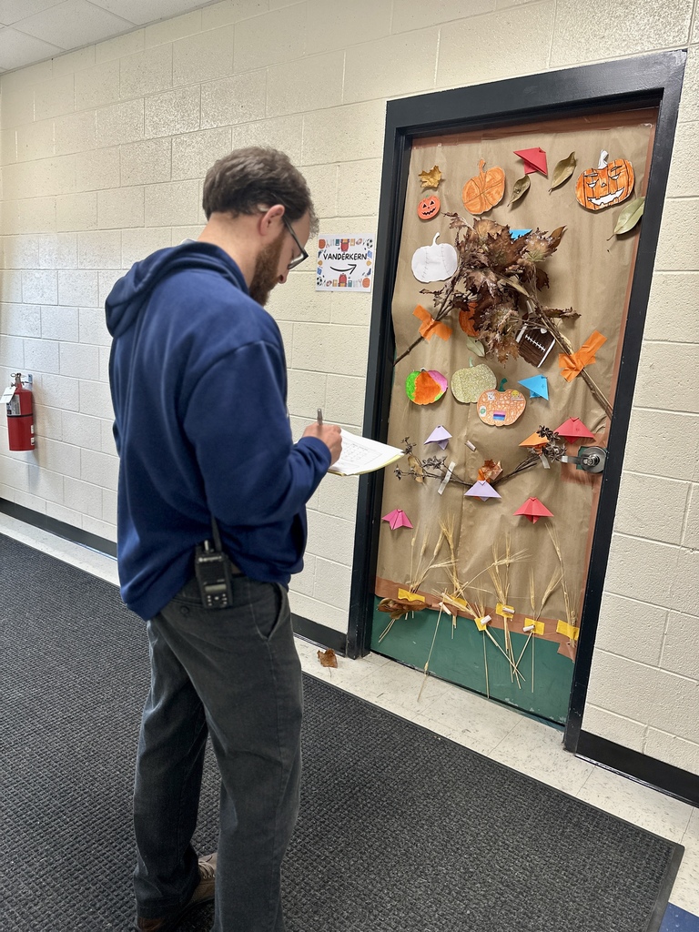 A man in a blue sweatshirt judges a fall-themed classroom door decorated with leaves, pumpkins, and straw.