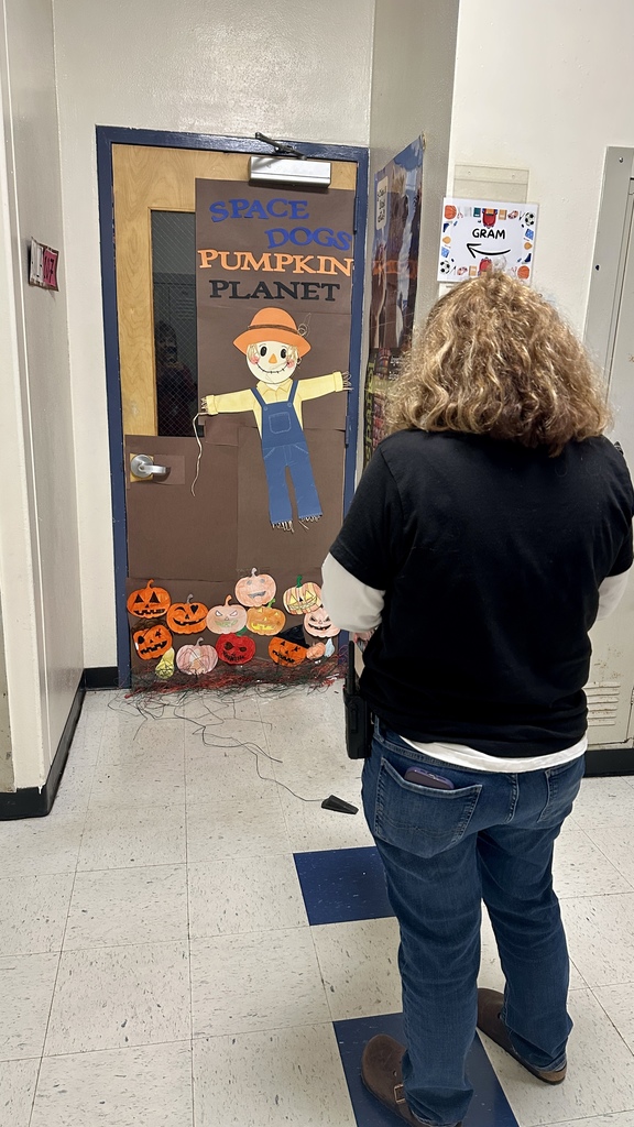 A person stands in front of a classroom door decorated with a scarecrow and pumpkins labeled “Space Dogs Pumpkin Planet.”