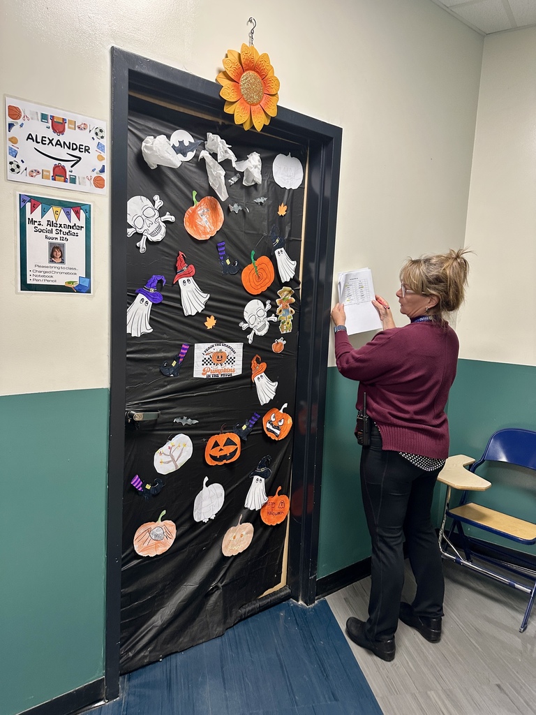 A woman with a clipboard judges a Halloween-themed classroom door covered in ghosts, pumpkins, and skulls.