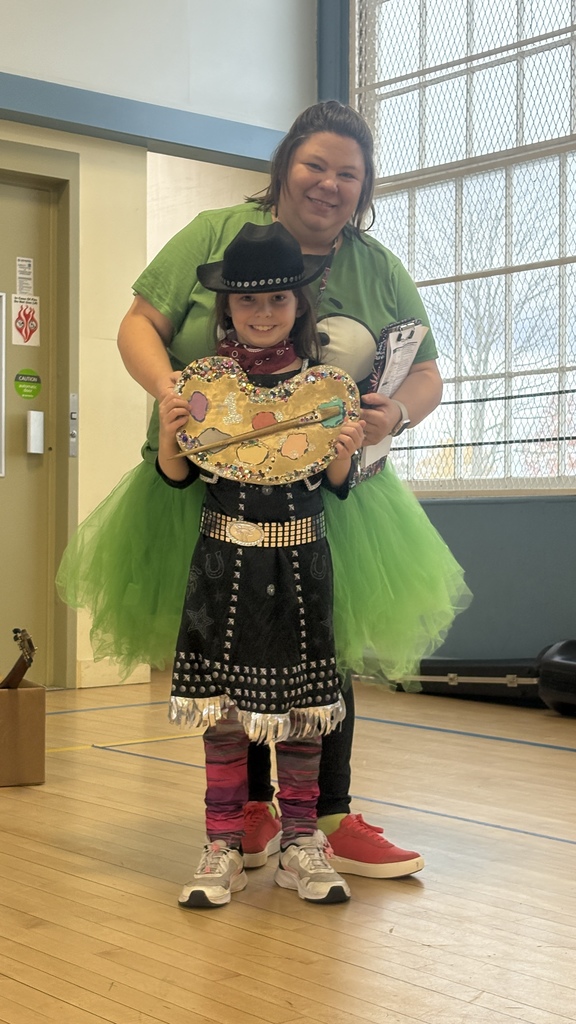 A smiling student wearing a black hat and western-style outfit holds a colorful art palette award, standing in front of an adult wearing a green shirt and green tutu, who is smiling and standing behind them during a school assembly.