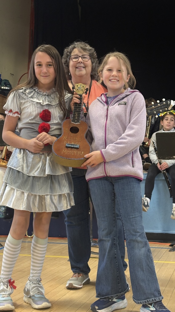 Two students stand with a music teacher holding a small decorated ukulele labeled “Class of the Month.” One student wears a silver costume dress with red pom-poms, while the other is dressed casually in jeans and a jacket.