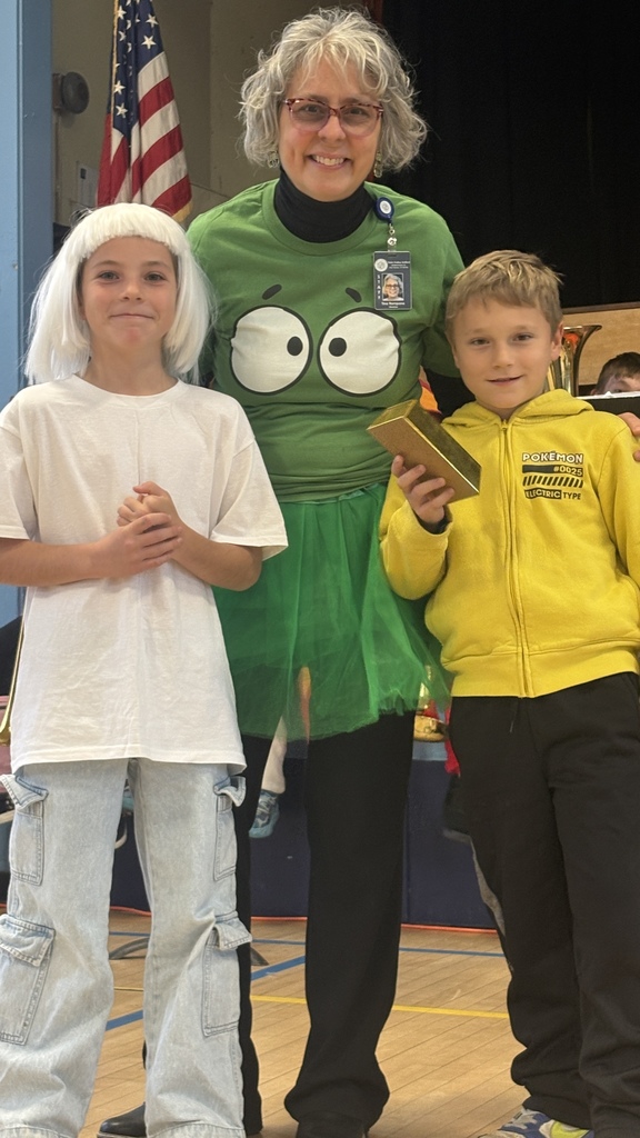 Two students stand beside a teacher wearing a green shirt and tutu. One student wears a white wig and shirt, while the other wears a yellow Pikachu hoodie and holds a gold brick prop. All three are smiling during a school event.