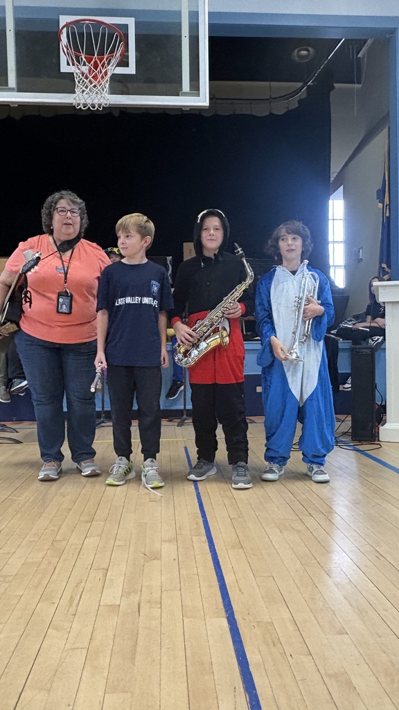 Three students stand on stage with their music teacher during an assembly. The teacher holds a guitar, and the students each hold instruments—a flute, saxophone, and trumpet. They are dressed in a mix of school attire and costumes.