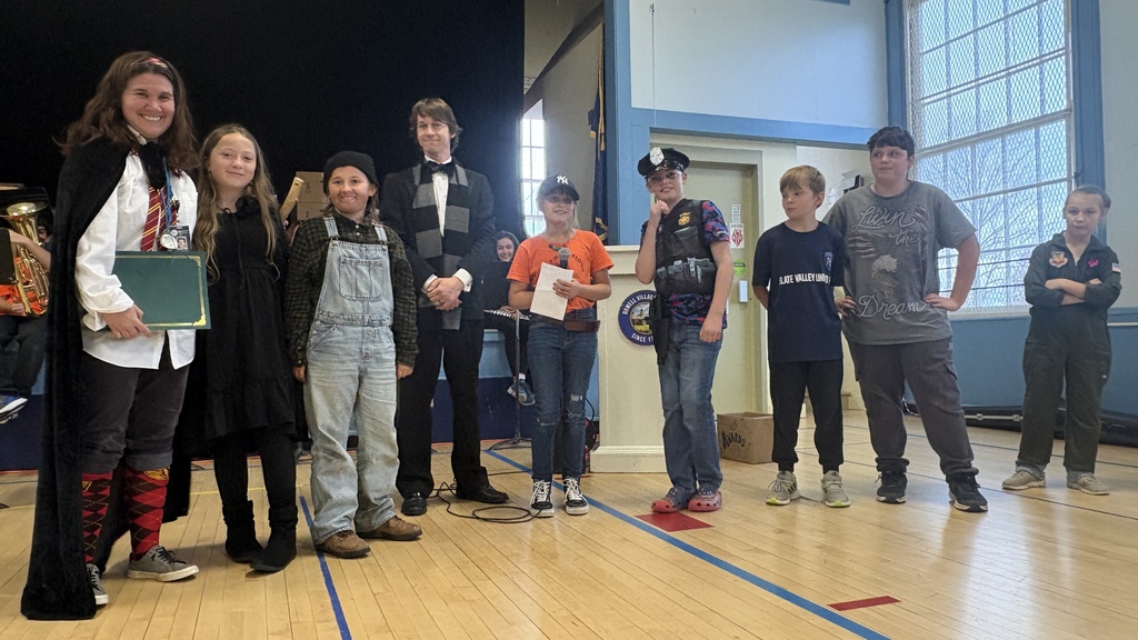 A group of students and teachers dressed in various costumes—such as a wizard, scarecrow, police officer, and pilot—stand on stage together during a school assembly, smiling and holding awards or props.