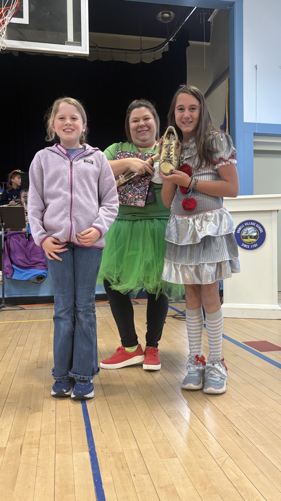 Two students stand with a teacher wearing a green tutu, smiling and holding a large golden shoe trophy as part of a school award ceremony.