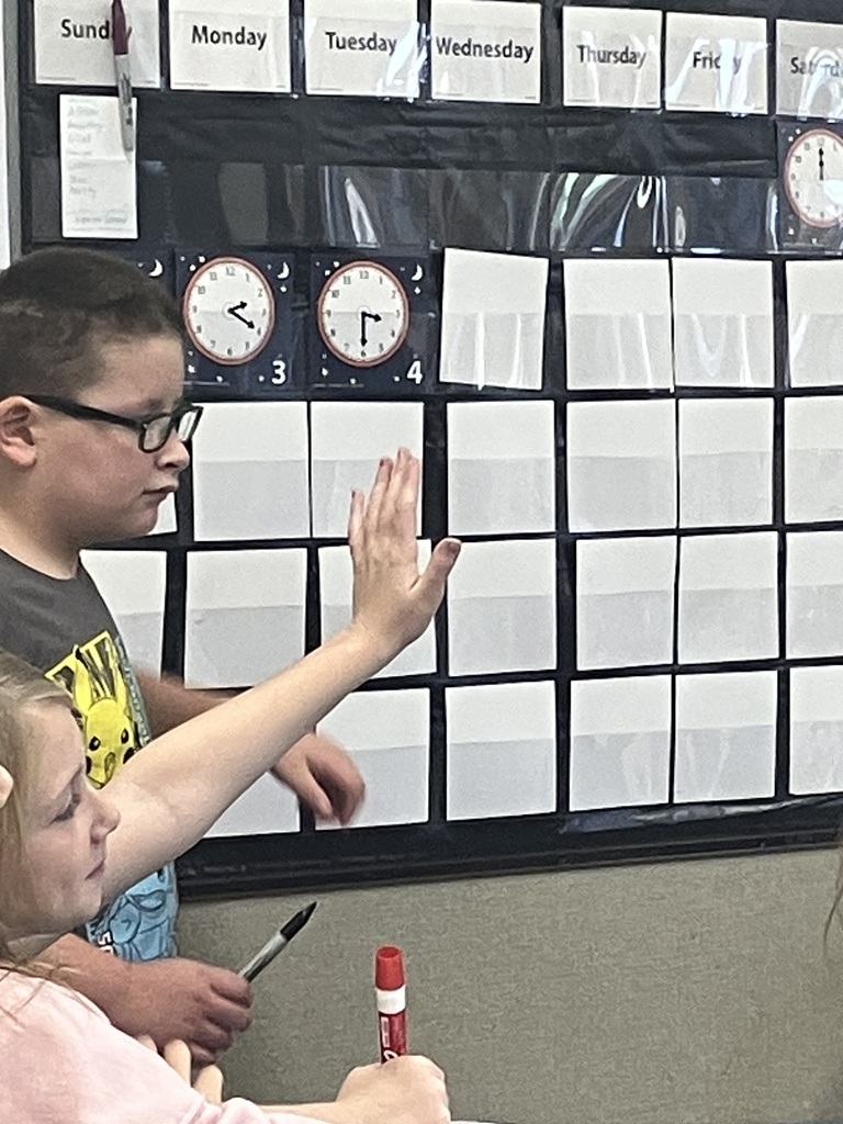 A boy wearing glasses is standing next to a calendar wall chart, pointing or raising his hand while a girl, visible in the foreground, is holding a red dry-erase marker. The calendar displays the days of the week, with two smaller charts underneath showing analog clocks with hands at different times, labeled '3' and '4'.  