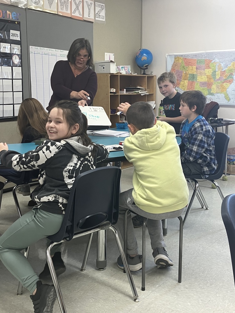  A group of elementary school-age children is seated around a blue table in a classroom. A female teacher, wearing a dark shirt and glasses, stands behind the table, addressing the students and pointing to a small whiteboard. A girl in a black and white hoodie is smiling brightly, while a boy in a neon yellow hoodie and two other boys look toward the teacher. A large map of the United States is visible on the wall behind them.  
