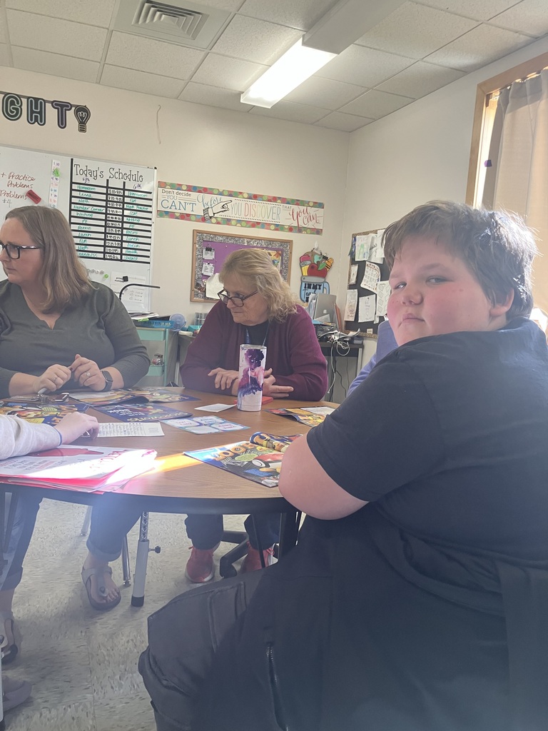 A group of people, including two adult women, are seated around a round wooden table in a brightly lit classroom. The woman on the left has glasses and long hair. The woman in the center is wearing a red sweater. A student in a black shirt is seated in the foreground, facing away from the table and looking directly at the camera with a neutral expression. An assignment board is visible on the wall behind them. 