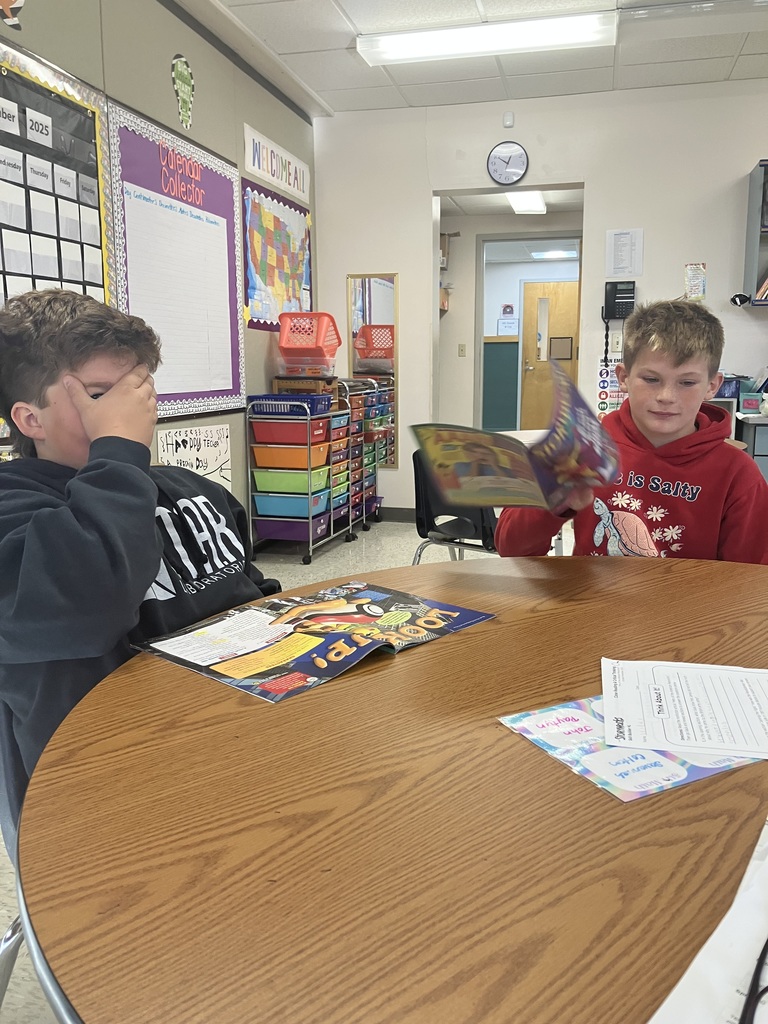   Two boys are seated opposite each other at a round wooden table. The boy on the left, wearing a black sweatshirt, has his hand covering his eyes as he looks down at an open magazine on the table. The boy on the right, wearing a red hoodie, is looking at and holding up his open book. Classroom decorations, including a calendar chart and colorful storage bins, are visible in the background.
