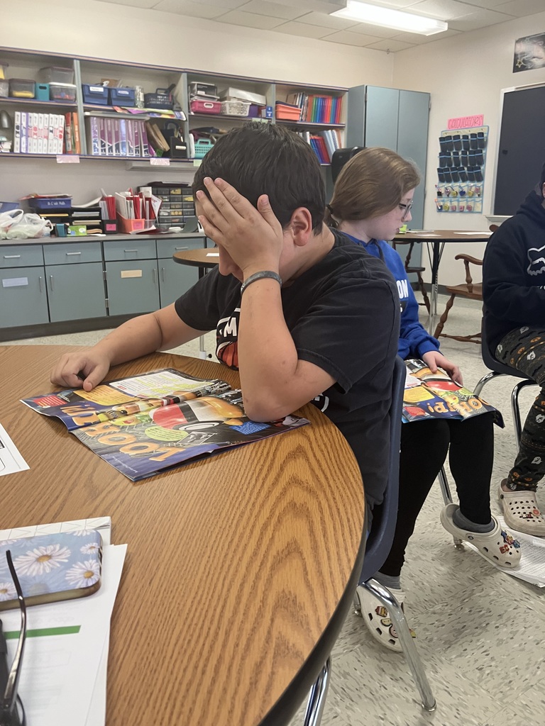 A close-up shot of a student seated at a wooden round table in a classroom. The student, wearing a black shirt, is looking down at an open book or magazine with colorful graphics, holding their hand up to their forehead. Another student is seated back-to-back behind them. Classroom shelves filled with organized supplies are visible in the background.  