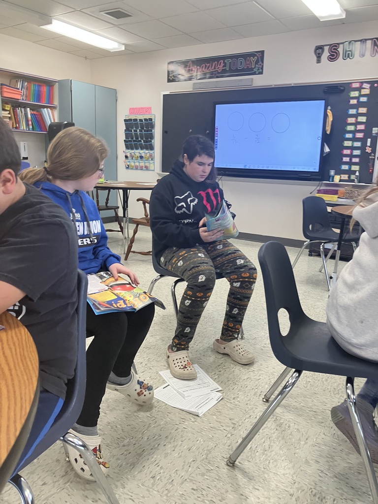  A student in a blue sweatshirt is seated at a table, looking down at an open book. Another student, wearing a dark hoodie and patterned pajama-style pants, is seated in a separate chair facing the front of the classroom and reading a book. In the background, a large interactive whiteboard displays three large blank circles, and classroom decor is visible on the walls.  