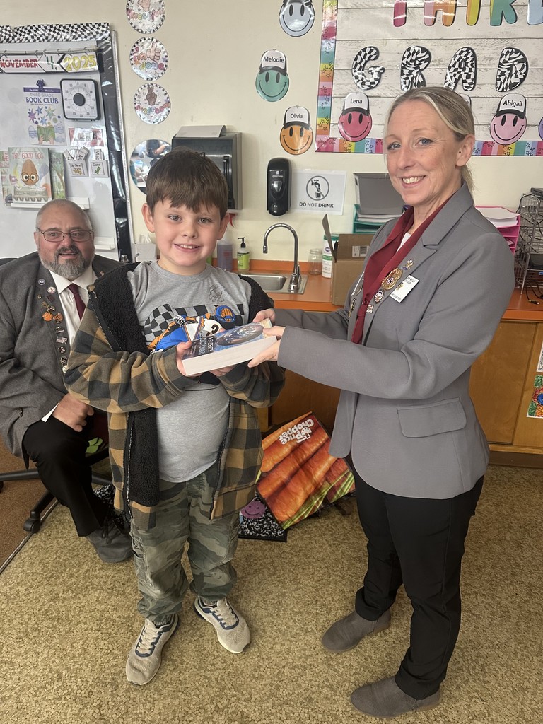  A woman in a gray blazer and black pants, wearing a pin, hands a book to a smiling young boy in a brown and black plaid jacket and camouflage pants. A man with a beard and glasses, wearing a dark suit and tie with a pin, sits in a chair behind the boy. The scene is a classroom with colorful decorations on the wall.