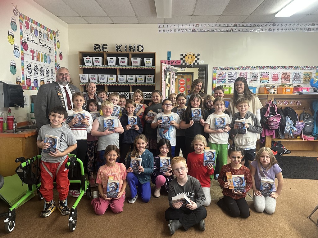  A group photo in a brightly decorated classroom shows a man and three women standing behind or slightly interspersed with about twenty young students. All the children are holding a copy of the same book, and the adults are smiling. A green mobility aid is visible on the left. The classroom wall has decorations like a "BE KIND" sign and happy face graphics.