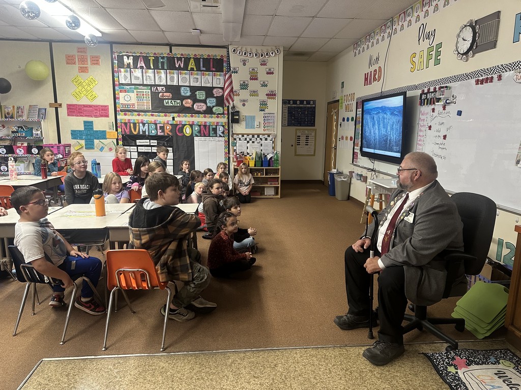  A man in a gray suit and tie sits in a chair facing a group of young students in a classroom. The students are seated at desks and on the floor, attentively looking toward the man. There is a TV screen on the wall displaying an image of a snowy forest. Classroom decorations include a "MATH WALL" and "NUMBER CORNER."
