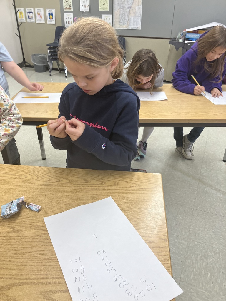  A girl in a dark blue "Champion" hoodie is seated at a wooden desk, focused on manipulating a small piece of wood or a broken pencil tip with her fingers. A piece of paper in front of her has a vertical column of numbers written on it, including 10, 20, 30, and up to 500. Other students are seated at desks in the background, writing.