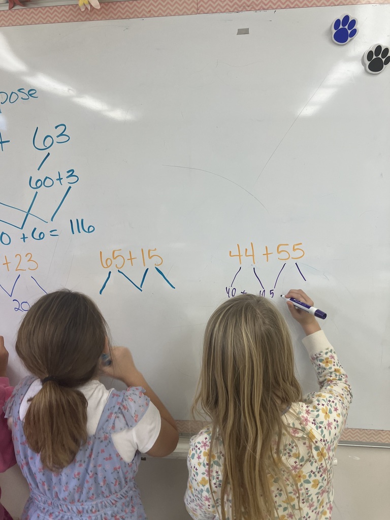  A view from behind two girls who are standing at a whiteboard, actively working on math problems. The girl on the right, with long blonde hair, is writing the steps to solve $44 + 55$ by decomposing the numbers. The girl on the left is wearing a blue floral dress. The board displays other decomposed addition problems