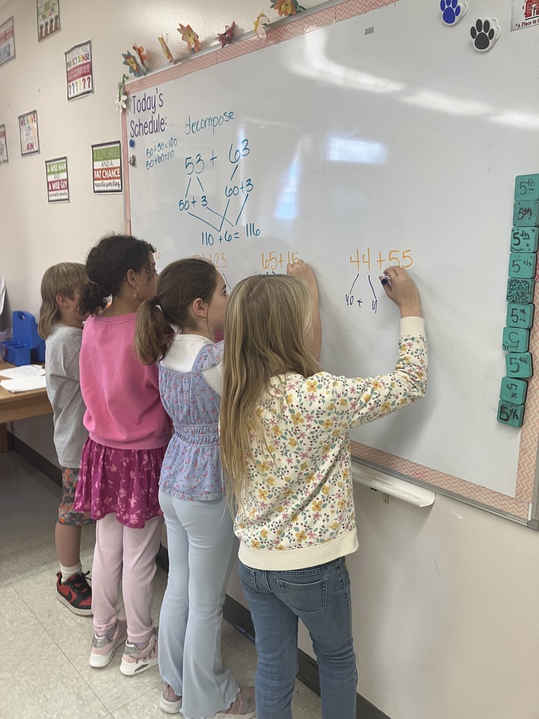  A wider shot showing four elementary school-age girls lined up at a whiteboard, all actively writing and solving addition problems using the number decomposition strategy. The whiteboard shows the math problem $44 + 55$ being worked on