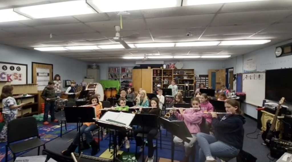 Elementary students play instruments together in a music classroom. Several students are seated with flutes, clarinets, and saxophones, while others play drums and percussion in the back. The room is brightly lit with posters, music stands, and instruments lining the walls.