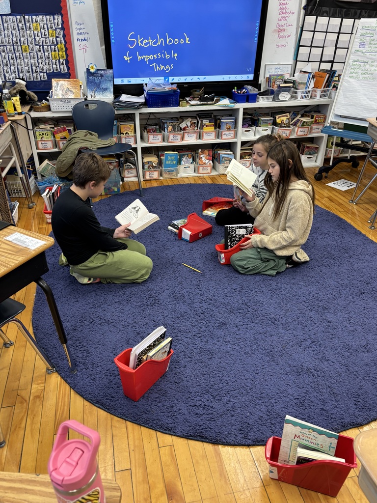 Three students sit on a blue rug reading books together in front of a classroom display labeled “Sketchbook of Impossible Things.”