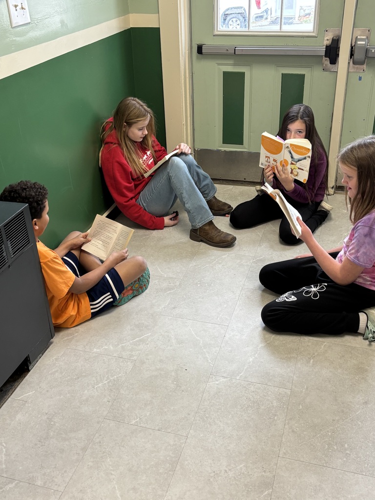 Four students sit in a hallway near a green and white wall, each reading a book quietly.