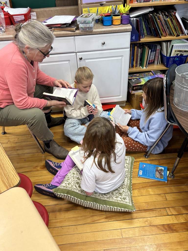 A teacher and three students sit on the floor in a reading group, each holding a book, surrounded by shelves of classroom materials.