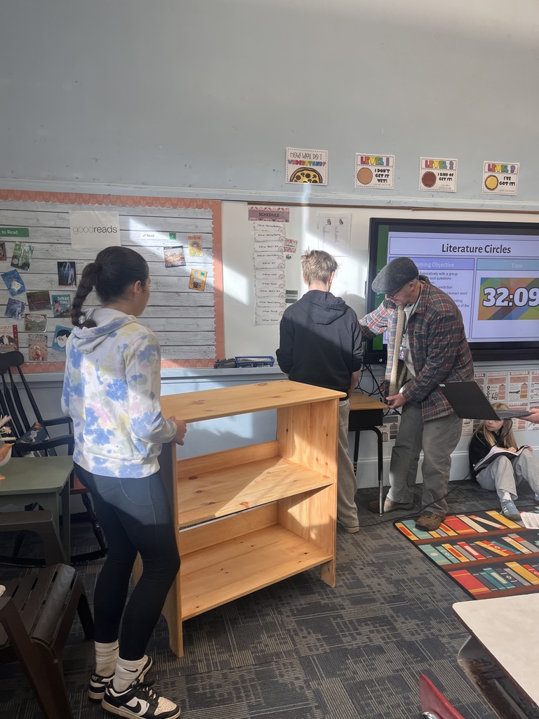 Two students and an adult adjusting a bookshelf in a classroom.