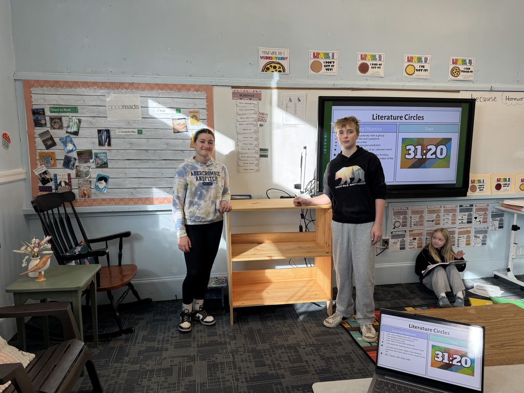 Two students standing next to a bookshelf in the front of a classroom.