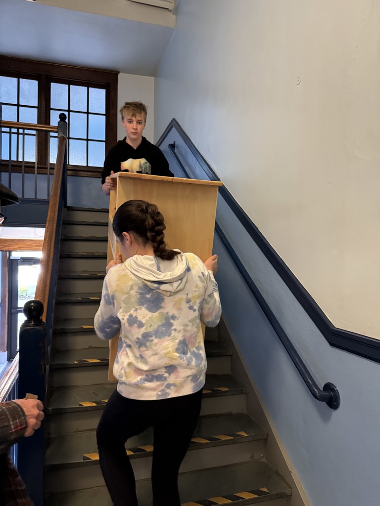 two students carrying a bookshelf up a flight of stairs.