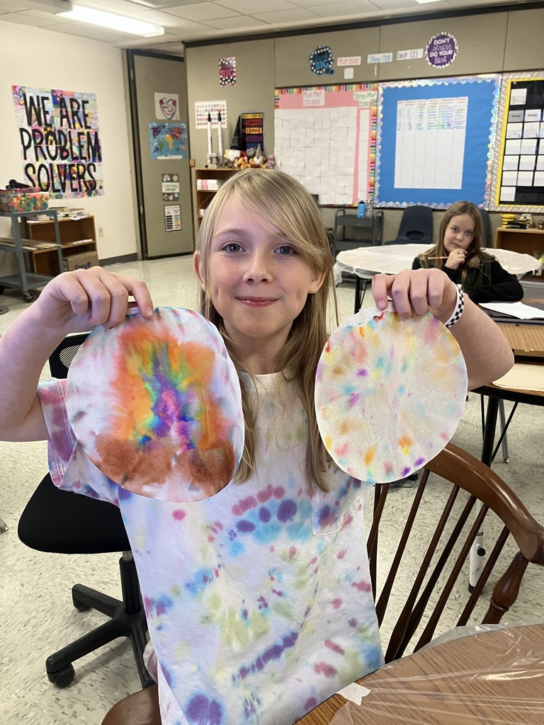 A girl with long blonde hair and a tie-dye shirt is seated, holding up two decorated coffee filters. The one on the left has bright rainbow colors, and the one on the right has lighter pastel colors. Another girl is visible seated at a table in the background. 