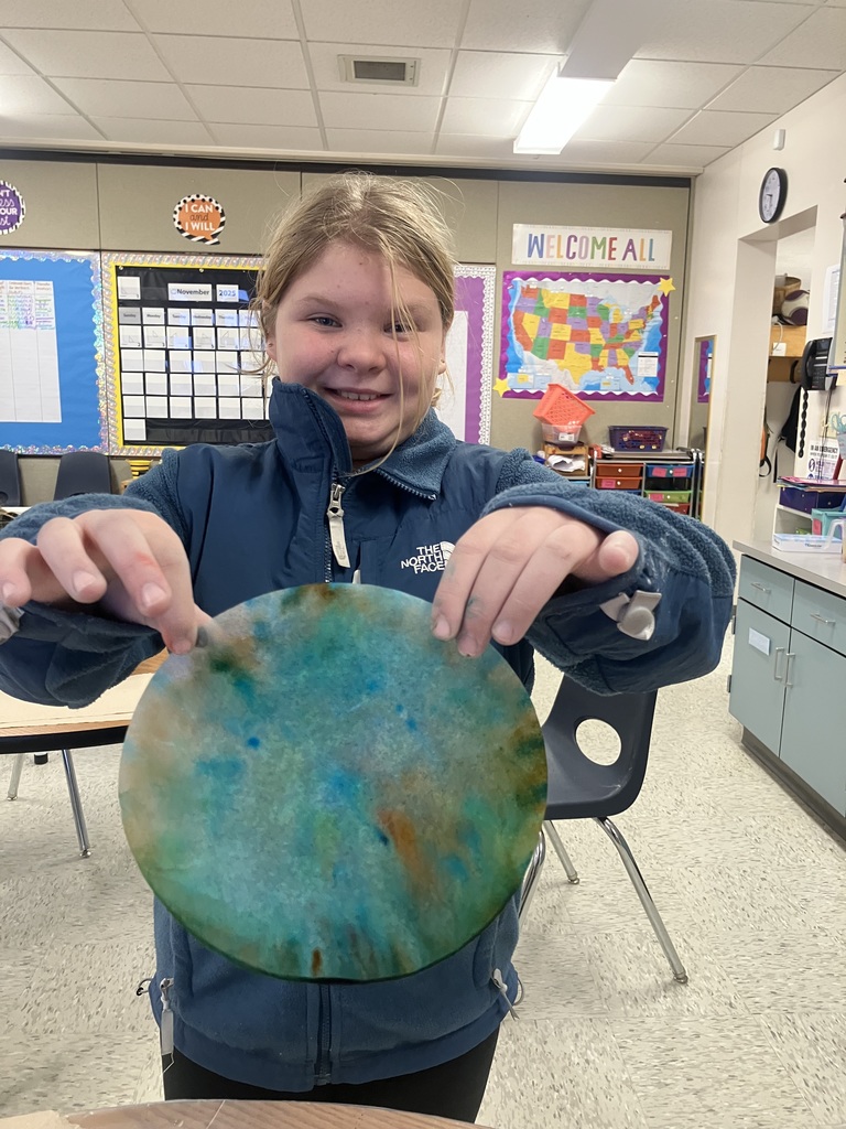  A smiling girl wearing a dark blue fleece jacket stands in a classroom, holding up a round piece of paper (likely a cniature Earth or tie-dye art. A cale USA are behind her.  