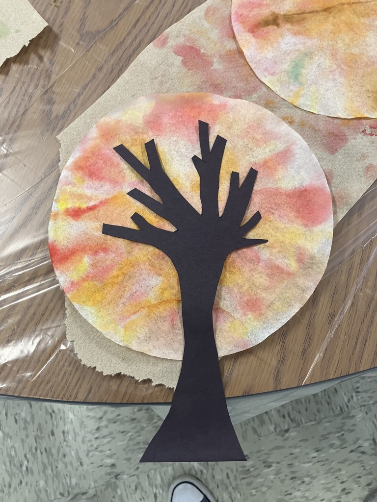 A close-up of a finished art project on a wooden tabletop covered in plastic. A black cutout silhouette of a bare tree is glued onto a round coffee filter dyed with red, orange, and yellow colors, giving it the appearance of an autumn sunset or fall foliage. 