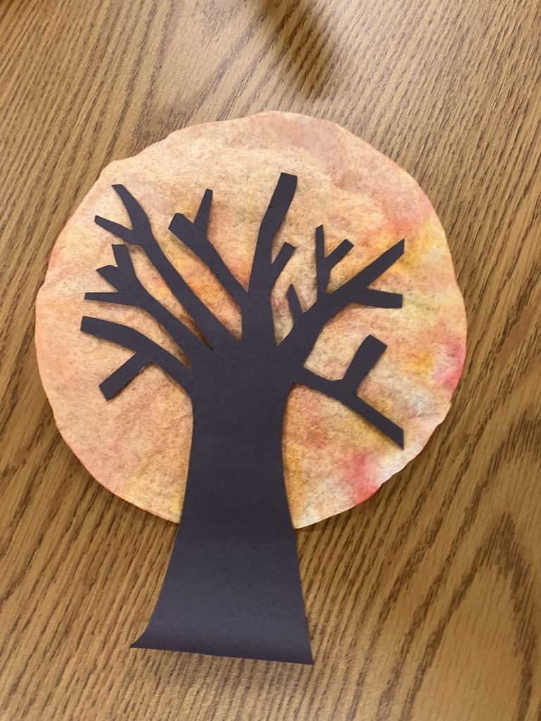  A close-up of a finished art project on a wooden tabletop covered in plastic. A black cutout silhouette of a bare tree is glued onto a round coffee filter dyed with red, orange, and yellow colors, giving it the appearance of an autumn sunset or fall foliage.  