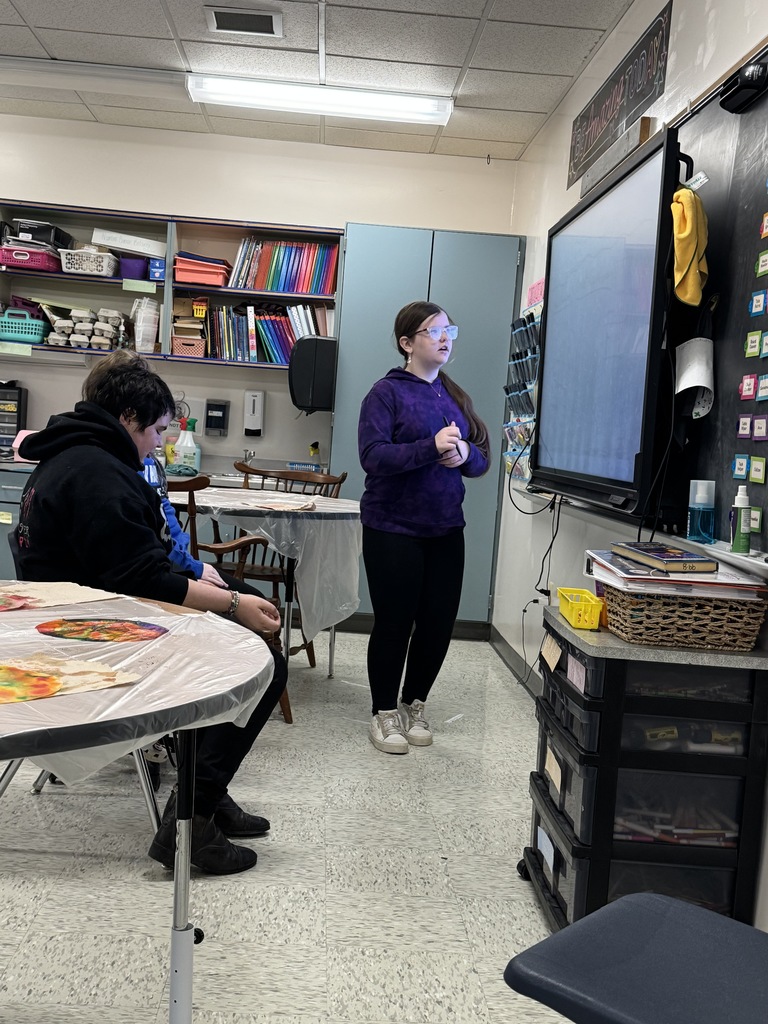  A classroom scene where a girl in a purple sweatshirt and glasses is standing by a large, dark interactive display screen, appearing to present or lead an activity. Another student is seated at a table covered in plastic on the left. Classroom shelves filled with art supplies are visible behind them.  