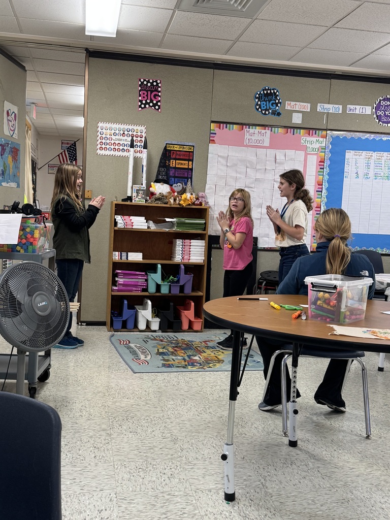  Four students are standing in a classroom. Three girls are facing a whiteboard with charts (labeled "Mat-Mat," "Stop-Mat"), and two of them appear to be singing or participating in a call-and-response activity, with their hands held near their mouths. The fourth girl is seated at a round table, facing away from the camera.