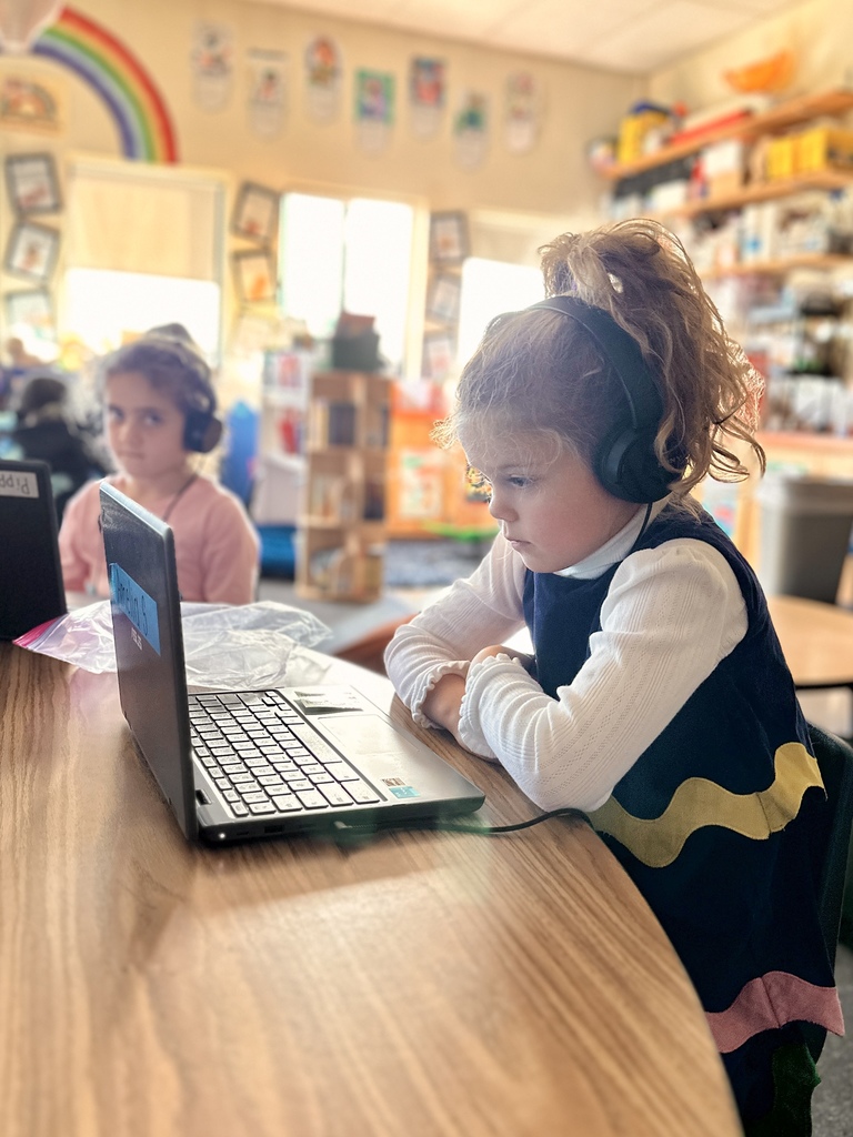 Student wearing headphones focuses on a laptop during classroom work; another student sits beside her.
