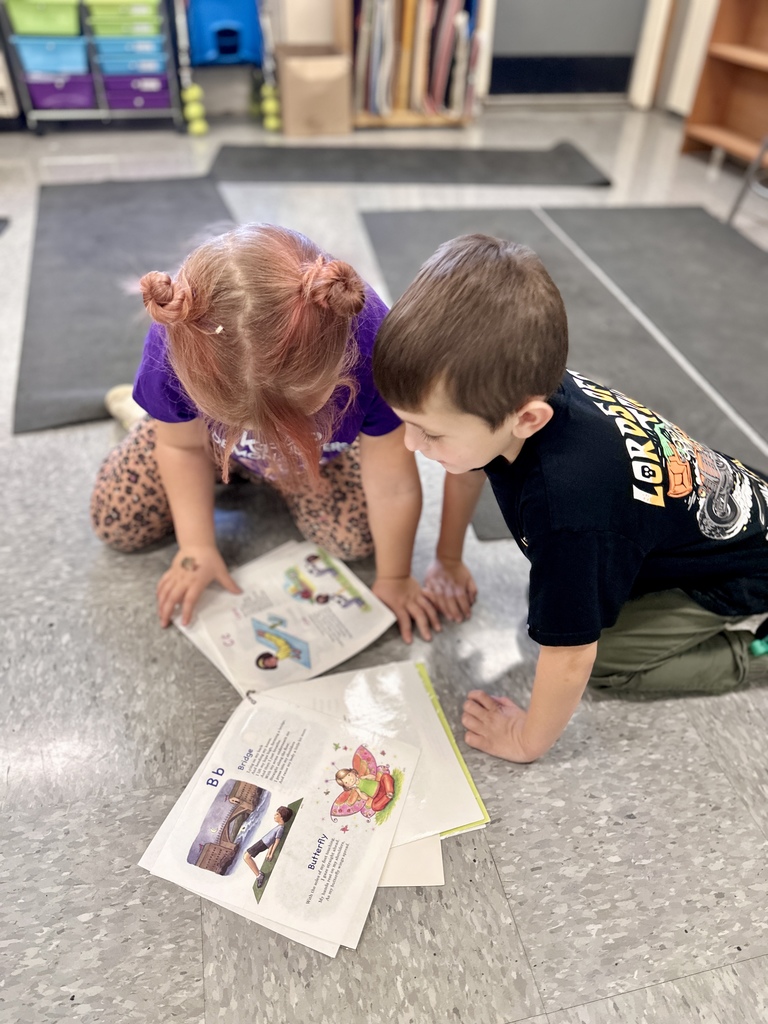 Two students kneel on the classroom floor and read a book together.