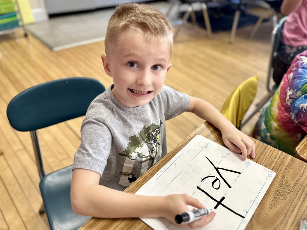 Young boy sitting at a classroom desk smiles while writing the word “yet” on a small whiteboard.