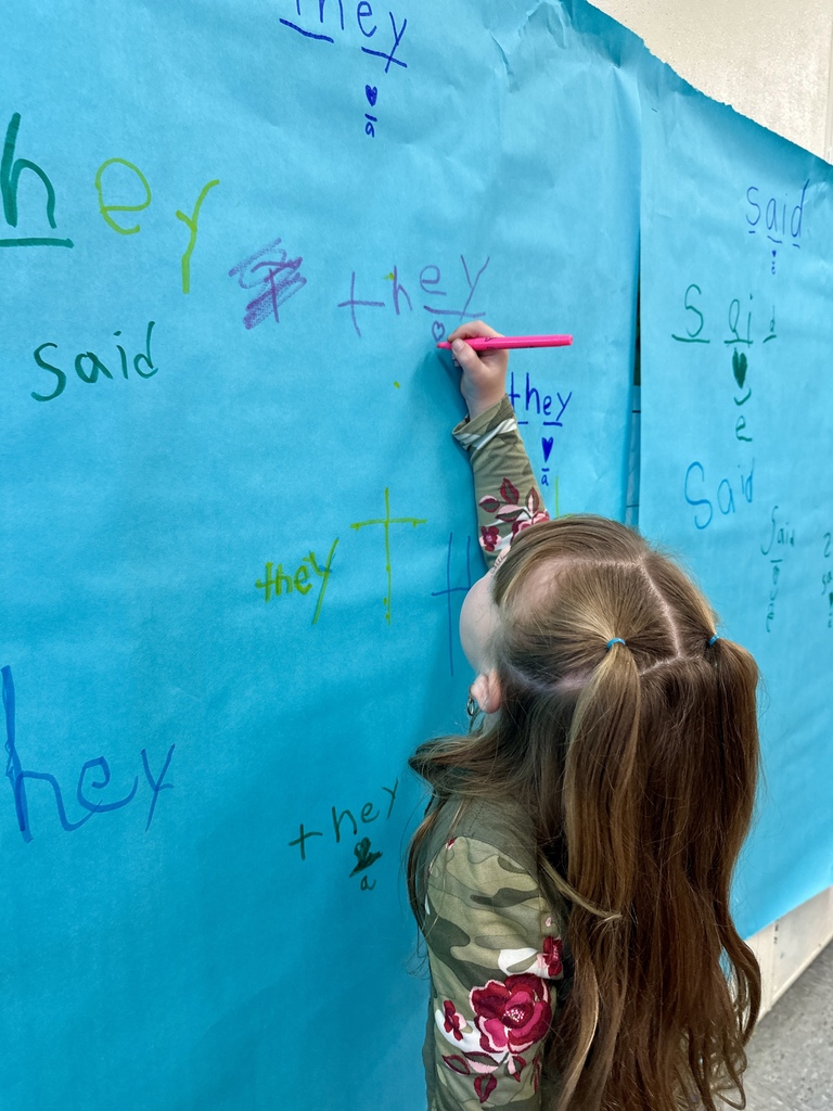 Girl writes the word “they” on large blue paper hanging on a classroom wall.