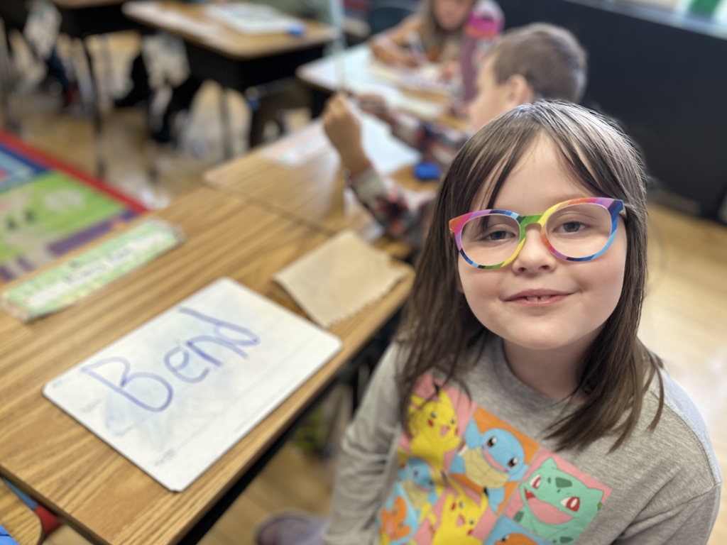 Girl wearing rainbow-colored glasses smiles at her desk with a whiteboard that has her name written on it.