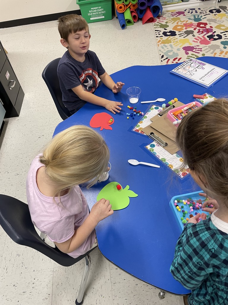 Three young students are seated around a blue, kidney-shaped table in a classroom, engaged in a craft activity. In the foreground, a girl with blonde hair in a ponytail is wearing a pink shirt and working on a green apple-shaped paper cutout, with small white pom-poms nearby. Another girl, with long brown hair and a green plaid shirt, is on the right, picking small colorful pom-poms from a blue tray. A boy in a dark t-shirt is at the top of the table, looking up and away from his work, which includes a red apple cutout, a small cup of white pom-poms, and a plastic spoon. Craft supplies like crayons and paper are also on the table.