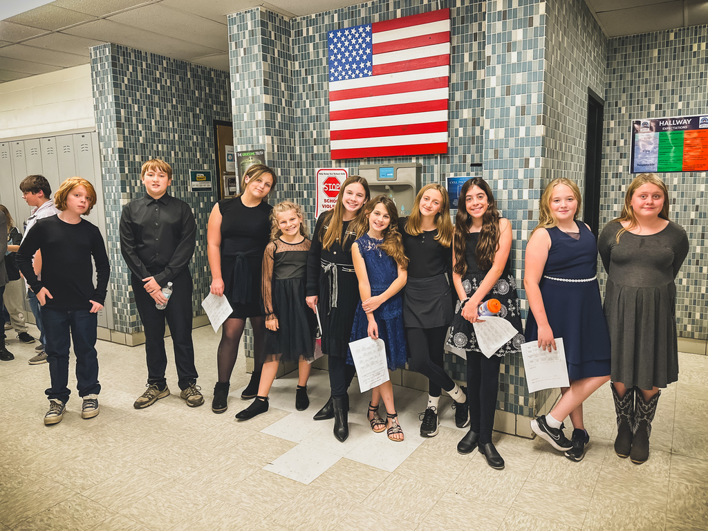 A group of students dressed in formal concert attire stand together in a school hallway, smiling and holding their music sheets. An American flag hangs on the tiled wall behind them.