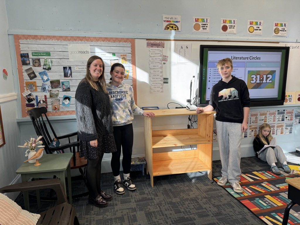 A teacher stands beside two students next to the newly delivered wooden bookshelf in a classroom. A third student sits on the floor reading near a colorful rug.
