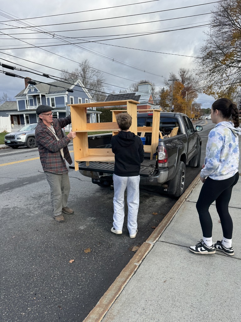 A man and two students stand behind a pickup truck parked along a street. They are unloading a wooden bookshelf from the truck bed. Houses and autumn trees line the background.