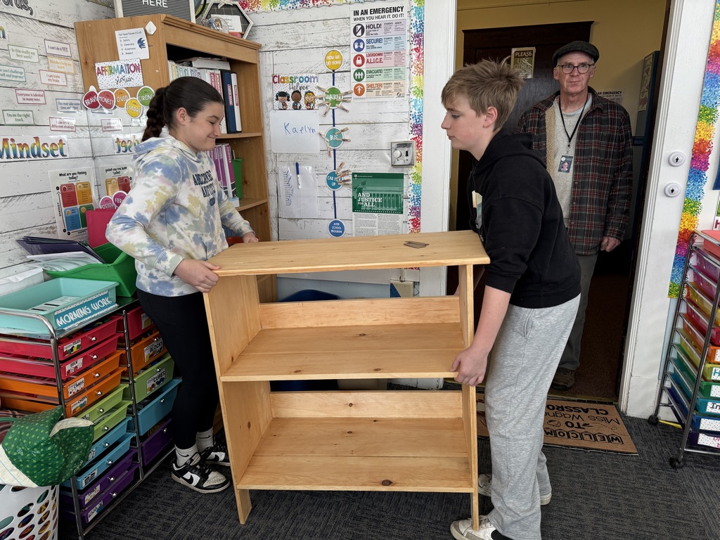 In a classroom, two students carefully place the wooden bookshelf on the floor while an adult stands in the doorway watching