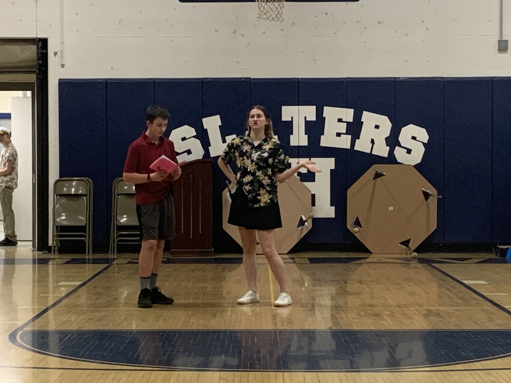 Two students stand in a school gymnasium rehearsing a scene. One student holds a script while the other gestures dramatically. Large letters spelling “SLATERS” are displayed on the wall behind them.